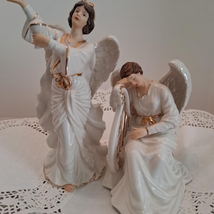 May include: Two white porcelain angel figurines. One angel is holding a chalice, the other is seated, leaning on a harp. Both angels have white wings, brown hair, and gold accents on their robes. The figurines are on a white lace doily.