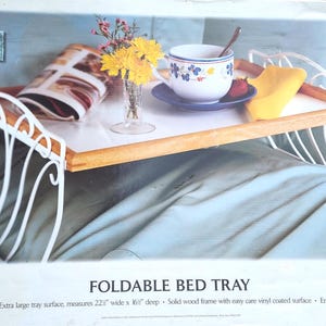 May include: A foldable bed tray with a solid wood frame and a white vinyl-coated surface. It features white enamel-coated decorative metal legs that fold. The tray measures 22.5 inches wide by 16 inches deep, as stated on the product packaging.