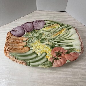 Puede incluir: Una fuente de cerámica con un diseño en relieve de varias verduras. La fuente presenta zanahorias, calabacines, tomates, maíz y cebollas en tonos de naranja, verde y rojo. El borde tiene una apariencia texturizada de tejido de cesta.