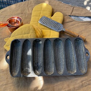 Puede incluir: Una sartén de hierro fundido para palitos de maíz con seis moldes en forma de mazorca, junto con guantes de horno amarillos, un rallador y tazas medidoras de cobre. Los artículos están dispuestos sobre una superficie de lino marrón, lo que sugiere un tema de cocina o repostería.