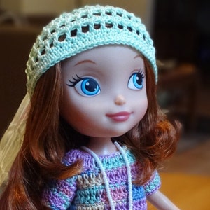 May include: A doll with long, curly red hair wearing a light blue crocheted hat and a multi-coloured crocheted dress with a white beaded necklace.