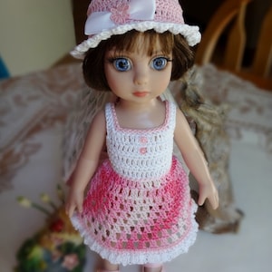 May include: A doll wearing a pink and white crocheted dress and a pink crocheted hat with a white ribbon. The doll has blue eyes and brown hair.