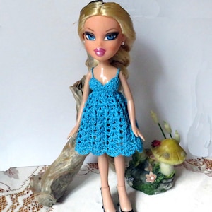 May include: A Bratz doll wearing a handmade, bright blue crochet dress with spaghetti straps. The doll has blonde hair, blue eyes, and is wearing black heels. The dress is a knee-length, open-weave design.