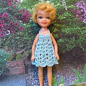 May include: A vintage doll with blonde hair and blue eyes wearing a light blue crocheted dress. The dress has a halter top and a flared skirt. The doll is standing in front of a background of flowers and greenery.