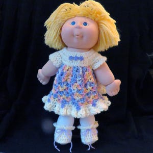 May include: A crocheted dress with a white ruffle and a lavender bow. The dress is worn by a Cabbage Patch doll with blonde hair and blue eyes.