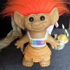 May include: A red-haired troll doll wearing a rainbow crocheted top and a crocheted orange skirt. The doll is holding a small mushroom with a ladybug on top.