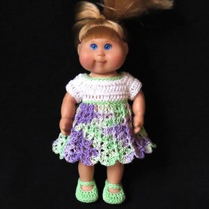 May include: A doll wearing a crocheted dress with a white bodice and a purple and green skirt. The doll has blonde hair and blue eyes and is wearing green crocheted shoes.