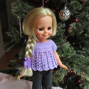 May include: A vintage doll with blonde hair styled in a braid, wearing a purple crocheted top and grey trousers. The doll is standing in front of a decorated Christmas tree.