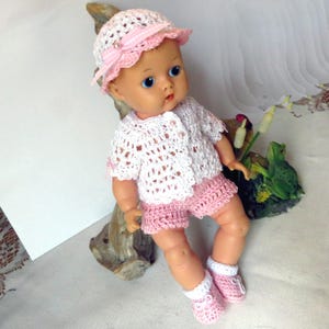 May include: A crocheted outfit for a baby doll, including a white and pink hat, a white cardigan with pink trim, pink shorts, and white and pink booties.