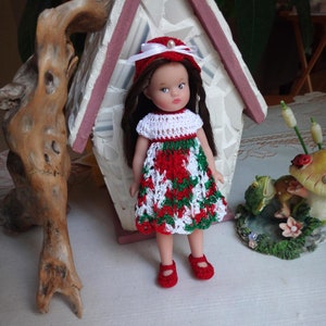 May include: A doll with long brown hair wearing a crocheted red, white, and green dress, a matching red crocheted hat with a white ribbon and a small pearl accent, and red crocheted shoes. The doll has a fair complexion and painted facial features.