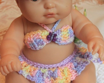 May include: A crocheted doll outfit in shades of purple, pink, yellow, and white. The outfit includes a bikini top with a butterfly embellishment, a skirt, and booties.