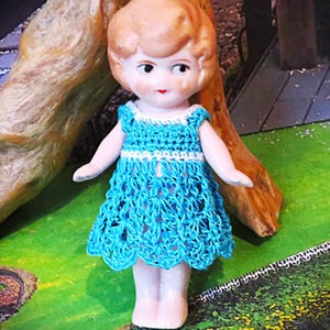 May include: A vintage porcelain doll with blonde hair and a blue crocheted dress. The doll has painted facial features, including rosy cheeks and red lips. The dress has a white trim around the neckline and sleeves. The doll is standing on a green surface.