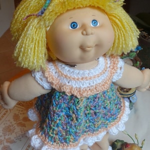 May include: A Cabbage Patch Kid doll with yellow yarn hair wearing a crocheted dress with a white lace trim. The dress is a mix of blue, green, pink, and peach colors.