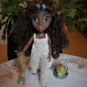 May include: A doll with dark brown curly hair and brown eyes wearing a white crocheted outfit. The doll is holding a piece of driftwood in her hand.