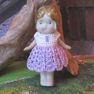May include: A vintage porcelain doll with blonde hair and a hand-crocheted dress. The dress features a white bodice and a lavender skirt. The doll has a painted face with delicate features and is posed against a natural wood background.