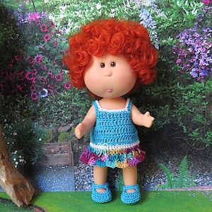May include: A handmade doll with bright red curly hair, wearing a blue crocheted dress with a rainbow-colored skirt and matching blue shoes. The doll has a fair complexion and is standing in front of a floral background.