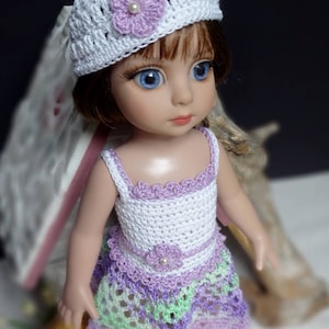 May include: A doll wearing a white crocheted hat with a lavender flower and a pearl center. The doll is also wearing a white and lavender crocheted dress with green accents. The doll has blue eyes and brown hair.