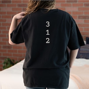 Puede incluir: Camiseta negra con los números blancos "312" impresos verticalmente. La camiseta es de cuello redondo y manga corta. La imagen muestra la parte trasera de la camiseta.