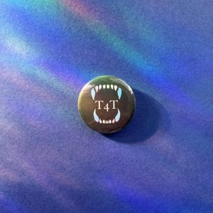 May include: A circular pin-back button featuring a black background with a graphic of an open mouth showing sharp teeth in shades of pink, white, and blue. The text T4T is printed in white in the centre of the mouth graphic on the button surface.
