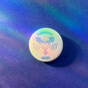 Puede incluir: Un botón circular con un fondo de degradado pastel. En el centro hay un símbolo en forma de corazón con alas y un halo, rodeado por el texto "TRANSSEXUAL ANGEL" en letras azules. El diseño incorpora símbolos de género.