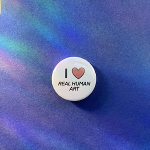 May include: A circular white pin-back button featuring a black capital letter I, a red heart symbol, and the black text REAL HUMAN ART printed in a sans-serif font. The design is centred on the button, which has a smooth, glossy finish.