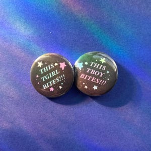 Peut inclure: Deux badges ronds à épingle avec un fond noir et une finition brillante. Le badge de gauche porte le texte THIS TGIRL BITES!!! en blanc, entouré de petites étoiles roses et blanches. Le badge de droite porte le texte THIS TBOY BITES!!! en blanc, entouré de petites étoiles bleues et blanches.