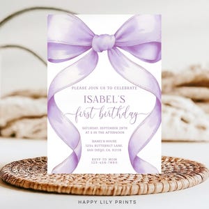 May include: A white invitation card with a watercolor-style lavender bow and ribbon design. The text reads "PLEASE JOIN US TO CELEBRATE ISABEL'S first birthday" with event details. The card is on a woven surface.