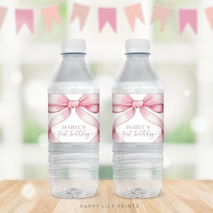 Może przedstawiać: Dwie przezroczyste plastikowe butelki z wodą z etykietami w postaci różowej wstążki i kokardki. Etykiety głosiły "Isabel's First Birthday".