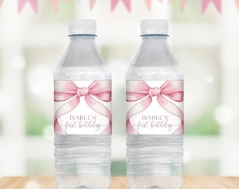 Puede incluir: Dos botellas de agua de plástico transparente con etiquetas de cinta rosa y lazo. Las etiquetas dicen "Isabel's First Birthday".
