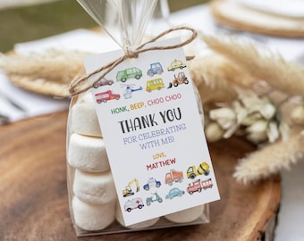 May include: A clear cellophane bag tied with twine, filled with marshmallows and a tag. The tag reads "HONK, BEEP, CHOO CHOO THANK YOU FOR CELEBRATING WITH ME! LOVE, MATTHEW" with vehicle illustrations. The bag sits on a wooden surface.