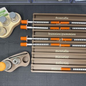 May include: An overhead shot shows medical supplies, including syringes with orange plungers and clear barrels, arranged on a gray surface. The syringes are labeled with anatomical terms. Also present are small bottles and a package of antiseptic wipes.