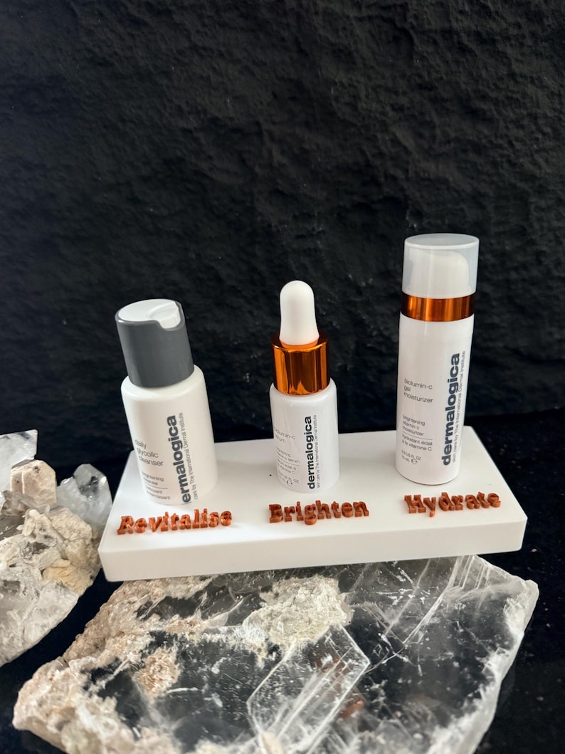 May include: Three white skincare product bottles with the brand name "dermalogica" on a white display stand. The stand has the words "Revitalise", "Brighten", and "Hydrate" in brown lettering. The bottles have silver and copper accents.
