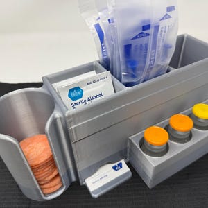 Può includere: Un organizer grigio stampato in 3D con più scomparti. Contiene pacchetti di alcol sterile, tamponi e flaconi con tappi arancioni e gialli. L'organizer ha anche un supporto a forma di tazza per i tamponi arancioni e una sezione per forniture mediche confezionate.