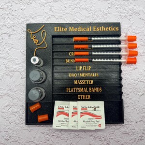 May include: A black organizer with the text "Elite Medical Esthetics" and labels for procedures. It holds syringes with orange caps, vials, and alcohol prep pads. The background is a textured white surface.