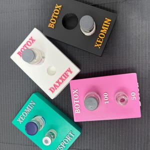 May include: Four coloured rectangular holders with text and vials. The holders are black, white, pink, and teal. The black holder has the text "Botox" and "Xeomin". The white holder has "Botox" and "Daxxify". The pink holder has "Botox", "100", and "50". The teal holder has "Xeomin" and "Dysport".