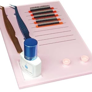 Może przedstawiać: Różowa taca do rzęs z przezroczystą butelką kleju do rzęs, dwoma pęsetami i paskami rzęs. Taca ma linie do organizowania rzęs i dwa małe kółka do trzymania kleju do rzęs.