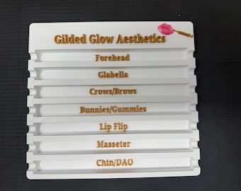 May include: A white plastic organizer with the text "Gilded Glow Aesthetics" in gold lettering. The organizer has multiple horizontal slots with labels including "Forehead," "Glabella," and "Crows/Brows."