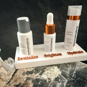 May include: Three white skincare product bottles with the brand name "dermalogica" are displayed on a white platform. The platform has the words "Revitalise", "Brighten", and "Hydrate" in brown lettering. The bottles are for a daily glycolic cleanser, vitamin-c serum, and moisturizer.