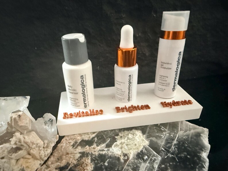May include: Three white skincare product bottles with the brand name "dermalogica" are displayed on a white platform. The platform has the words "Revitalise", "Brighten", and "Hydrate" in brown lettering. The bottles are for a daily glycolic cleanser, vitamin-c serum, and moisturizer.