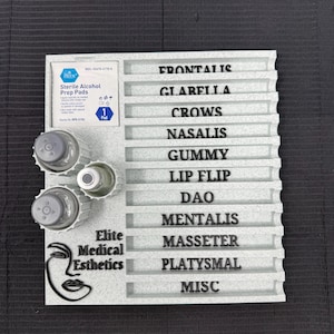 May include: A light gray esthetics tray with the text "Elite Medical Esthetics" and a stylized face logo. The tray has labeled slots for procedures like "Frontalis" and "Gummy", and includes alcohol prep pads and small bottles.