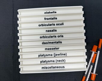 May include: A white plastic tray with labeled sections for facial anatomy terms like "alabella" and "frontalis." Three syringes with orange plungers are positioned to the right. The tray is on a dark surface.