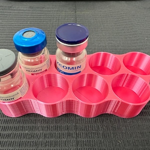 May include: A pink 3D-printed organiser holds several small glass vials with blue and grey caps. The vials contain a clear liquid and are labelled with the brand name "XEOMIN". The organiser has eight circular compartments.