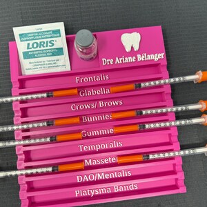 May include: A vibrant pink tray with labelled compartments for syringes. Labels include "Frontalis," "Glabella," and "Crows/Brows." Also visible are a small bottle, antiseptic pads, and the text "Dre Ariane Bélanger."