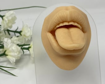 Puede incluir: Primer plano de una boca de silicona realista con la lengua fuera. La boca está abierta y muestra los dientes. La silicona es de un tono de piel claro.