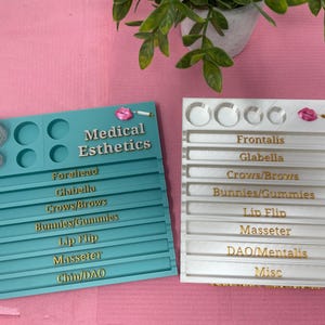 May include: Two trays, one teal and one white, with compartments for vials and labels. The teal tray has the words "Medical Esthetics" and lists areas of the face, such as "Forehead", "Glabella", "Crows/Brows", "Bunnies/Gummies", "Lip Flip", "Masseter", and "Chin/DAO". The white tray has the words "Frontalis", "Glabella", "Crows/Brows", "Bunnies/Gummies", "Lip Flip", "Masseter", "DAO/Mentalis", and "Misc".