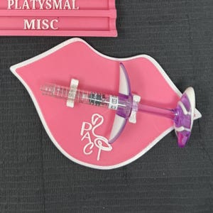 May include: A pink, lip-shaped tray with a white border holds a clear syringe with purple and white accents. The tray has the letters "PAC" and a flamingo design. Above, a pink shelf reads "PLATYSMAL" and "MISC".