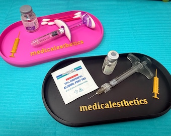 May include: Two oval trays, one pink and one black, with medical supplies. The pink tray has a syringe, cotton swabs, a vial, and a 3D-printed syringe. The black tray has a syringe, a vial, a 70% isopropyl alcohol prep pad, and a 3D-printed syringe. The text "medicalesthetics" is on both trays.