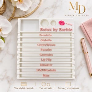 May include: A white rectangular organizer tray featuring nine horizontal channels labeled in pink text: Botox by Barbie, Frontalis, Glabella, Crows/Brows, Bunnies, Gummies, Lip Flip, Masseter, DAO/Mentalis, and Misc. Includes two circular vial wells.