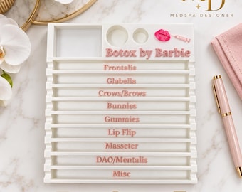 May include: A white rectangular organizer tray featuring nine horizontal channels labeled in pink text: Botox by Barbie, Frontalis, Glabella, Crows/Brows, Bunnies, Gummies, Lip Flip, Masseter, DAO/Mentalis, and Misc. Includes two circular vial wells.