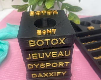 Puede incluir: Organizador de plástico negro con letras doradas. El organizador tiene seis compartimentos, cada uno etiquetado con un tipo diferente de inyectable: Botox, Jeuveau, Dysport, Daxxify y Xeomin.